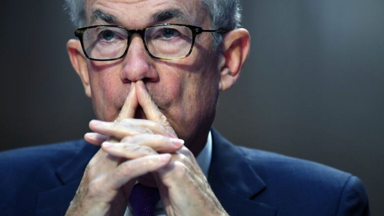 A man with grey hair and glasses holds his hands in front of his face at a press conference.