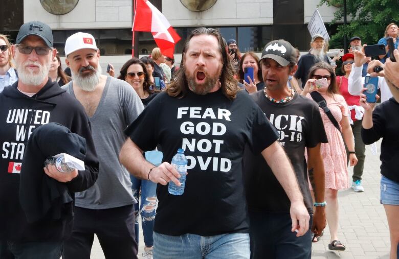 A crowd of people leave a courthouse in late spring. At the front is someone in a black t-shirt that says 'Fear God Not COVID.'