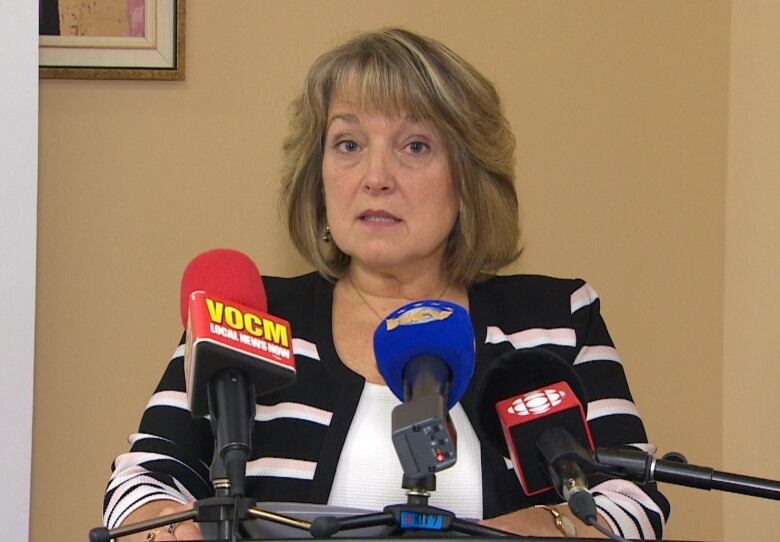 A woman stand at a podium with three microphones. The background is a beige wall. 