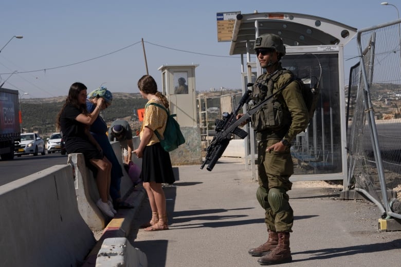 An Israeli soldier, in uniform and carrying a weapon, guards a bus stop while Israeli settlers wait for a ride at the Gush Etzion junction, the transportation hub for a number of West Bank Jewish settlements, Thursday, June 9, 2022.