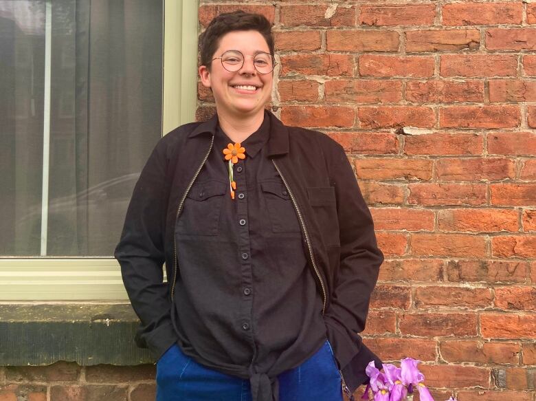 A person with short dark hair and glasses smiles while standing in front of a brick wall. 