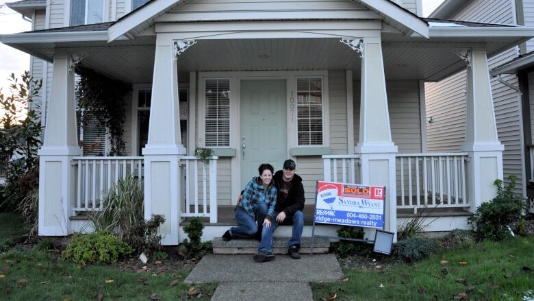 A couple sit on the porch of a home. Next to them is a for sale sign with a sticker that says 'sold.'