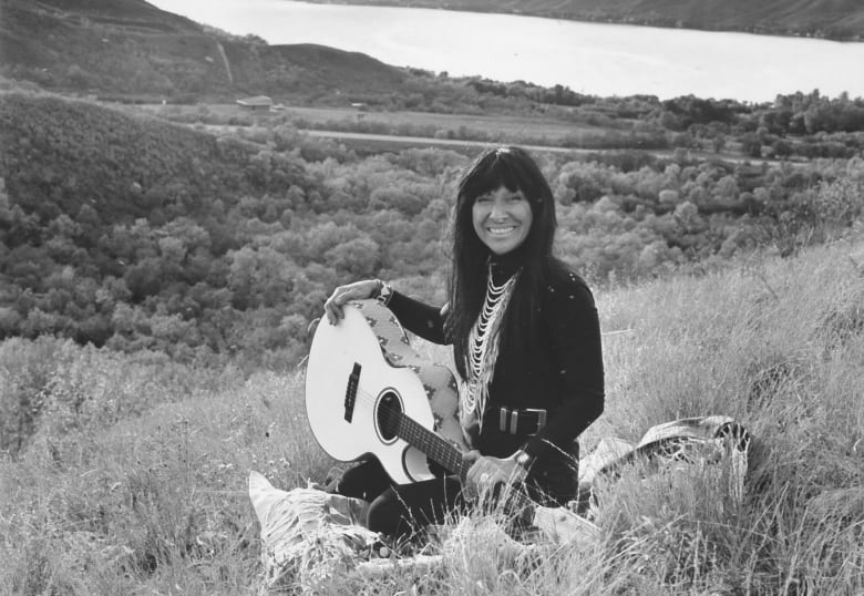 A woman sits in a grassy valley with a guitar.