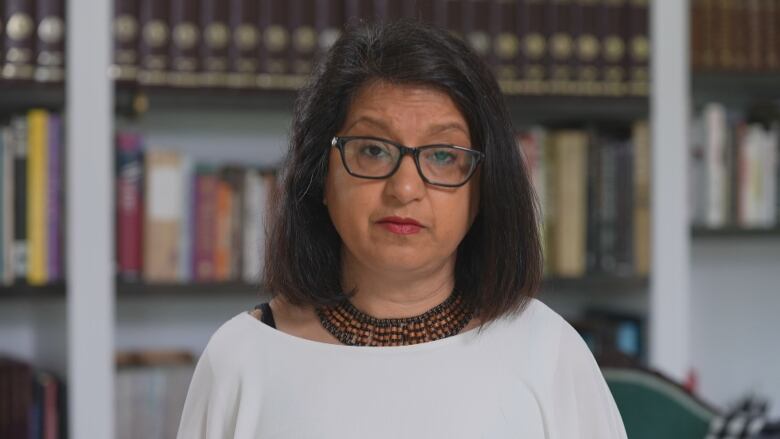 A woman wearing a white shirt and glasses stares ahead, with a bookshelf blurred in the background.