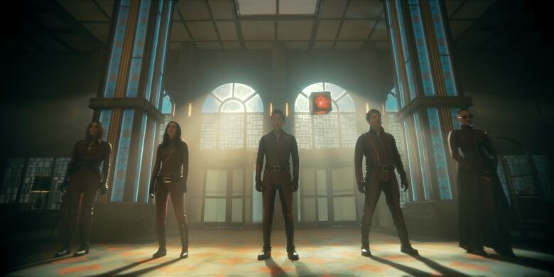 From left, five figures wearing red jumpsuits stand silhouetted against large windows in an empty room. 