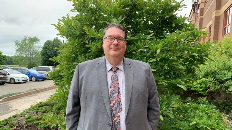 A man in a suit and tie standing in front of a green shrub