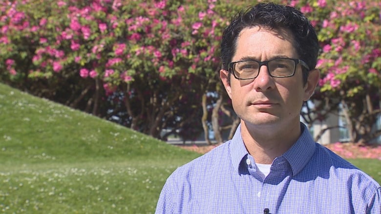 A man in an open-collar blue cotton shirt stands in front of cherry blossoms in a park.