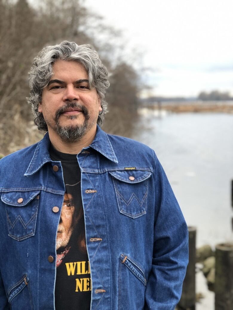 Daytime photo of the artist Brian Jungen, a middle aged man with wavy salt-and-pepper hair and a goatee. He stands in front of a body of water and wears a denim jacket and black Willie Nelson T-shirt.