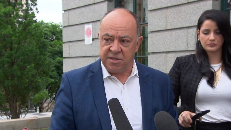 Quebec Health Minister Christian Dub, wearing a blue blazer, speaks to reporters outside in Quebec City, with a press aid to his left.