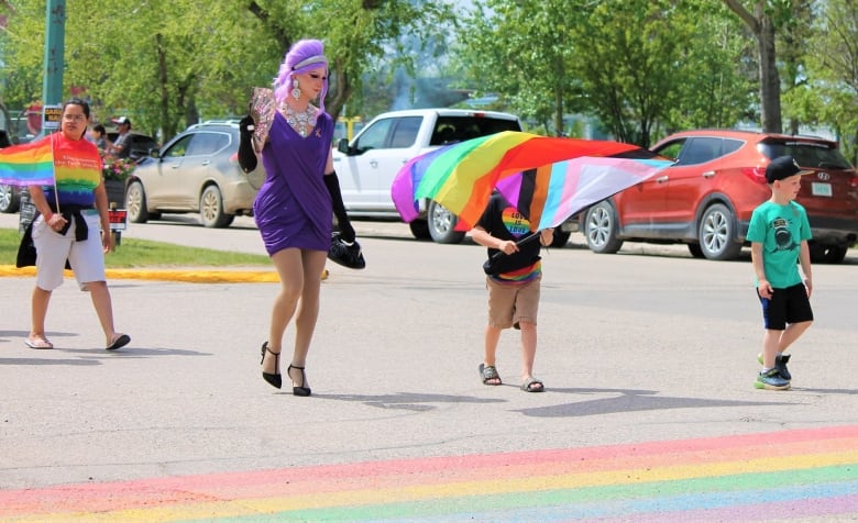 Four people are pictured crossing a rainbow crosswalk. Two are children, one is a woman and the last is in drag.