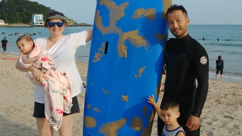 A woman with a baby in her arms, a man in a surf bodysuit and a young boy stand together near a surf board on a beach.