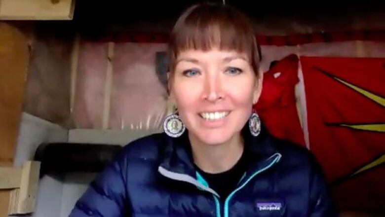 A woman smiles at the camera on a video call from inside a woodframe structure. 