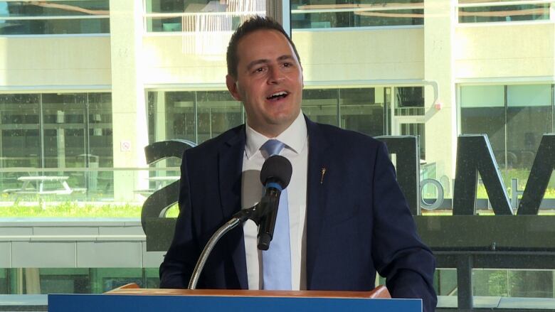 A man in a blue suit is seen talking with the help of a mic.