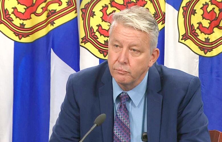 A man in a dark blue suit, light blue shirt and dark blue tie sits in front of a Nova Scotia flag.