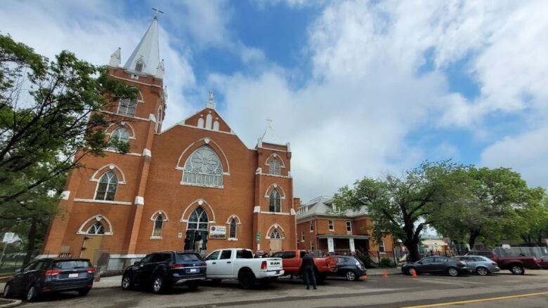 A brick church with large steeple stands above a city street lined with vehicles.