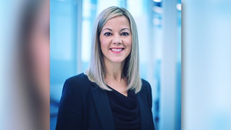 A person with straight, shoulder length blonde hair wearing a black blazer standing in front of a blue blurry background.