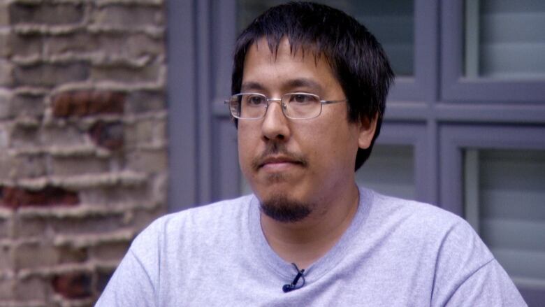 A man wearing glasses and a grey T-shirt sits outside.