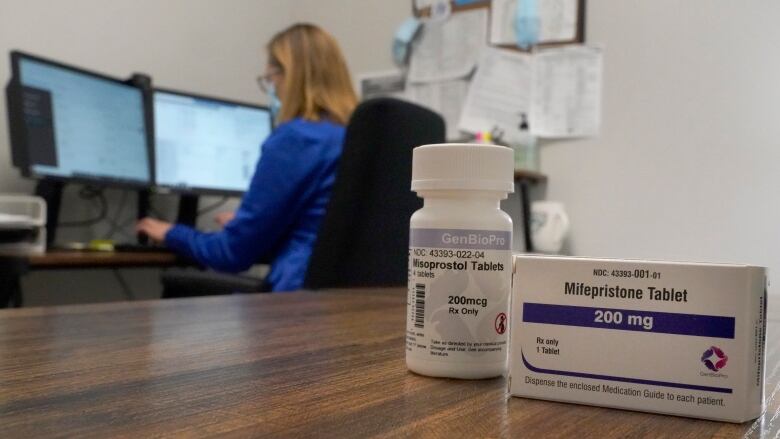 A box of Mifepristone tablets is in the foreground. A woman working at a computer in the background.