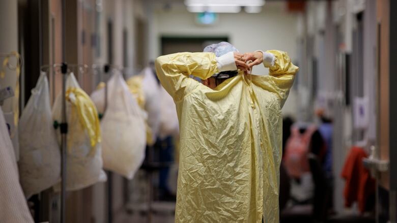 A healthcare worker standing in a hospital hallway, back to the camera, tying up a yellow protective gown.