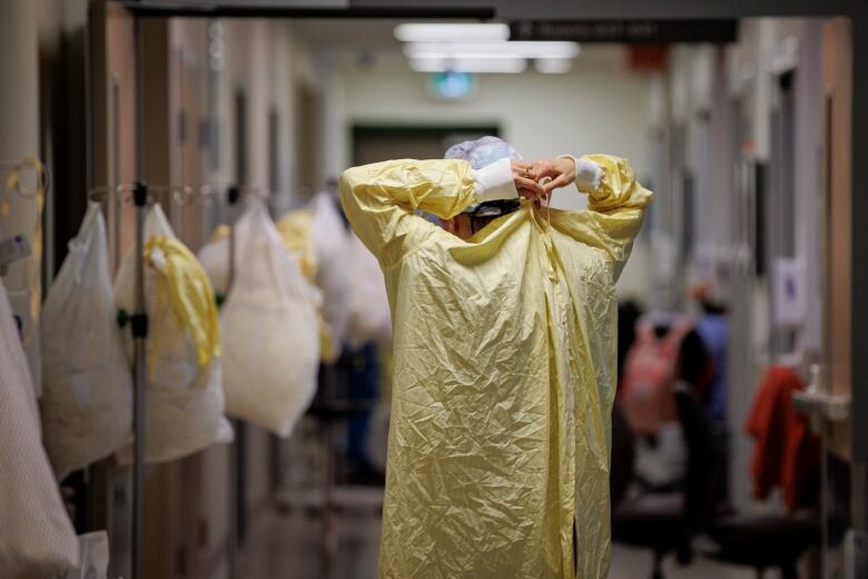 A healthcare worker standing in a hospital hallway, back to the camera, tying up a yellow protective gown.