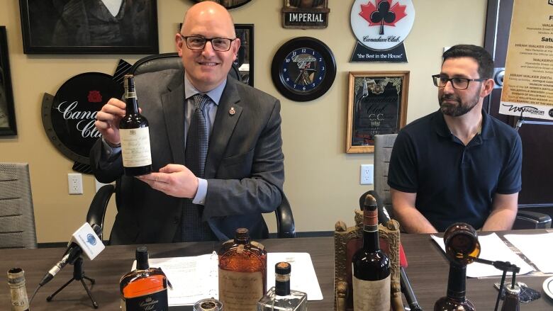 Two men are seated at a table covered with whisky bottles and memorabilia. 