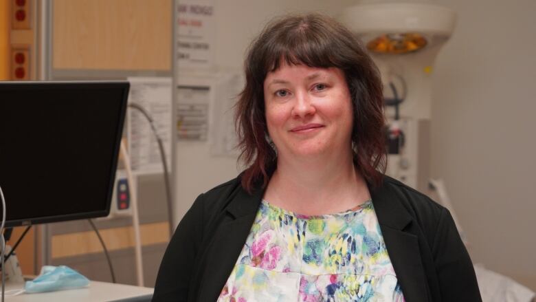 A white woman wearing a multicolour shirt and black cardigan standing inside a hospital room smiles at the camera.