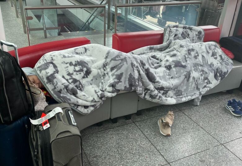 A woman is sleeping under a blanket, stretched over several airport chairs with luggage nearby.