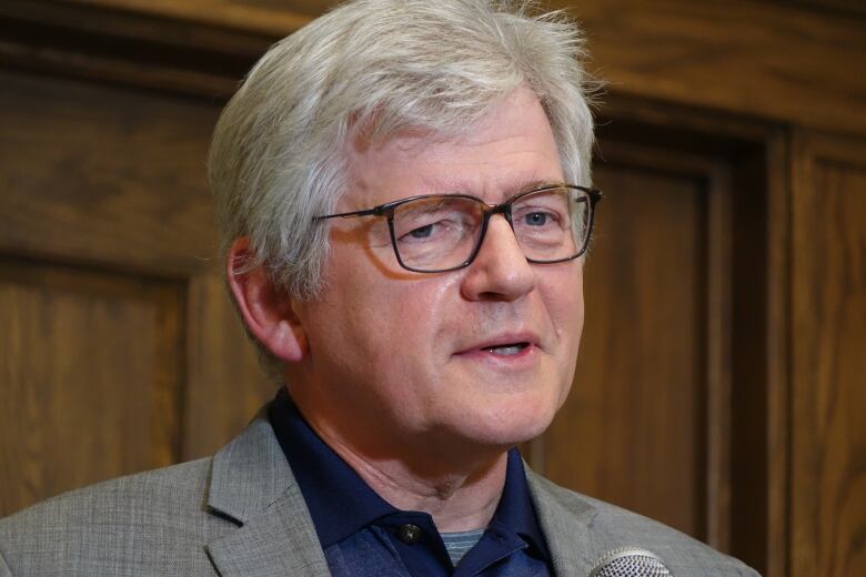 Jim Dinn stands in the media scrum area of the House of Assembly. He wears a grey blazer with a blue polo shirt, and has thin, brown glasses.