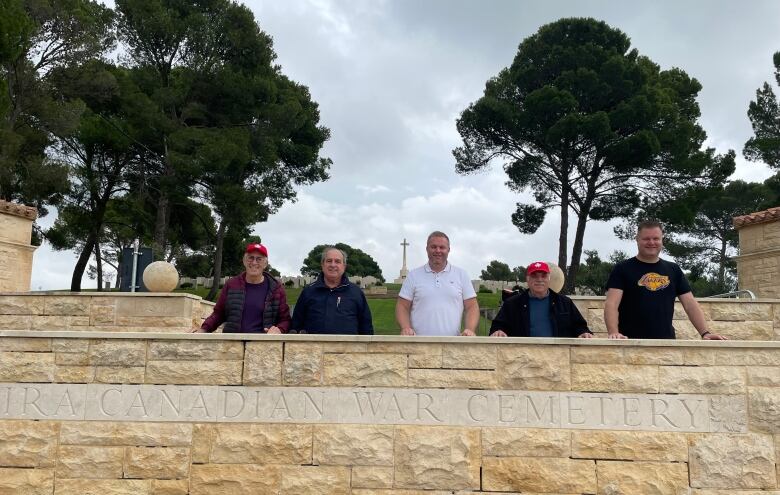 Five men standing behind a yellow stone wall. In the background is the cross monument in Agira, Sicily.