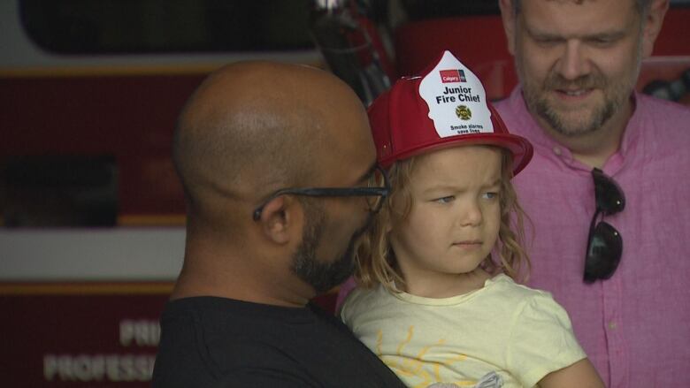 A man with a beard and glasses holds a little girl wearing a red hat that says 