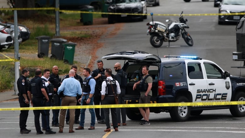 A group of men, many in uniform, stand at the back of a large police SUV. The men are encircled by crime tape. A police bike and police cars are visible in the background.