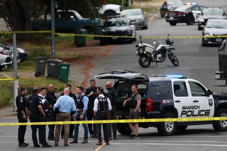 A group of men, many in uniform, stand at the back of a large police SUV. The men are encircled by crime tape. A police bike and police cars are visible in the background.