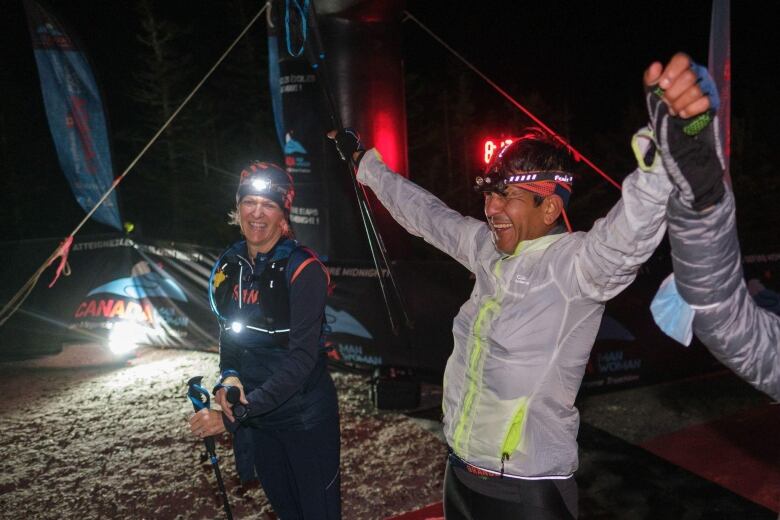 A man throws up his arms in victory, holding the hand of an unseen person. Another tired-looking runner, a woman, looks on.