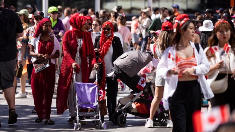 A large group of people, most of whom are wearing red to commemorate Canada Day. In focus are a family of three hijabi women. They all have red hijabs, and one of them is pushing a purple pushcart.