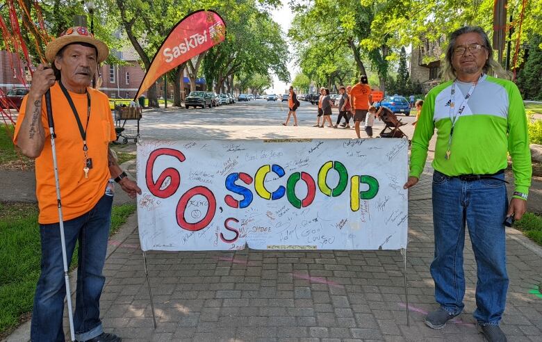 Two men stand in a park with a banner between them reading '60s Scoop' in large, colourful lettering, with many names written on it.