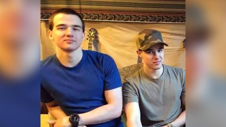 Two young men sit in front of a wall hanging, with a guitar neck visible in the background. The man on the left is wearing a blue shirt and has a black watch. The man on the right is wearing a grey shirt and has a camo hat.