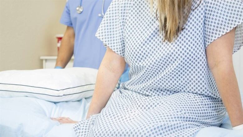 A woman wearing a medical gown sits on an examination bed as a health-care provider wearing medical scrubs and gloves, with stethoscope around their neck, enters the room.