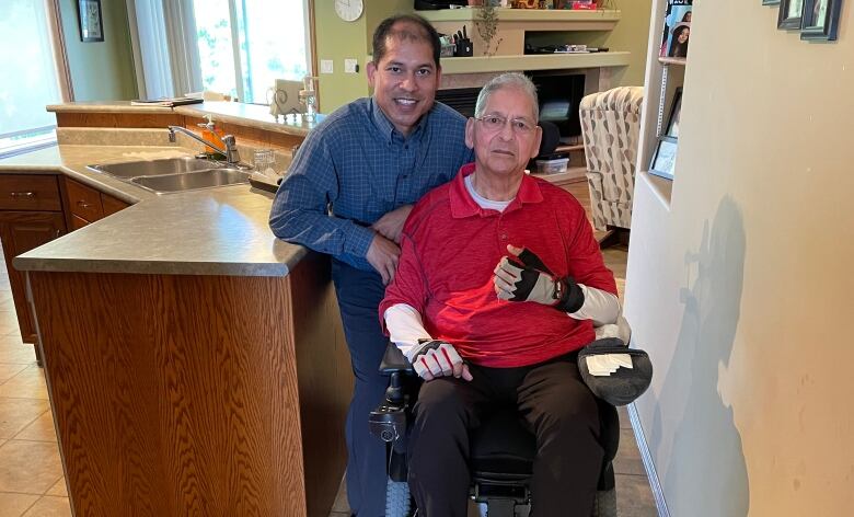 A senior is seated in a wheelchair, with a younger man standing behind him.