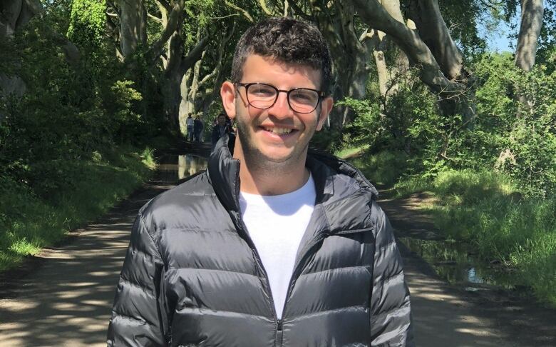 A man with glasses and a black puffer jacket smiles in front of trees.