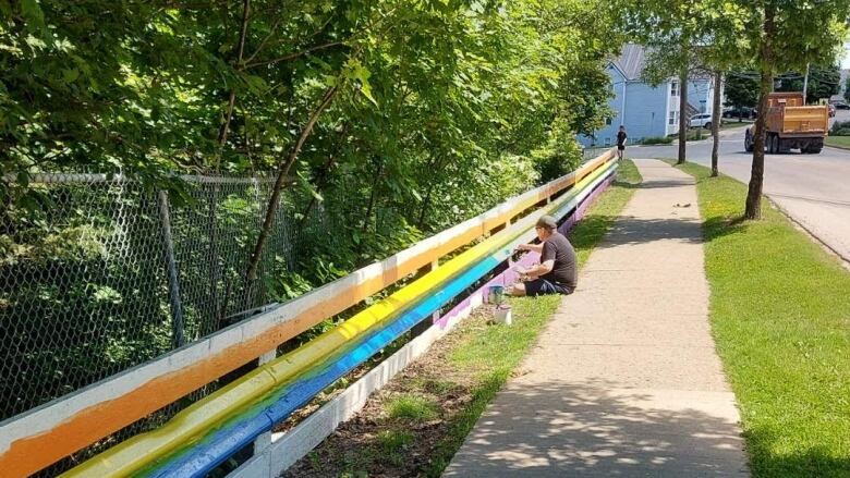 A person is seen sitting on a sidewalk painting a fence in rainbow colours.