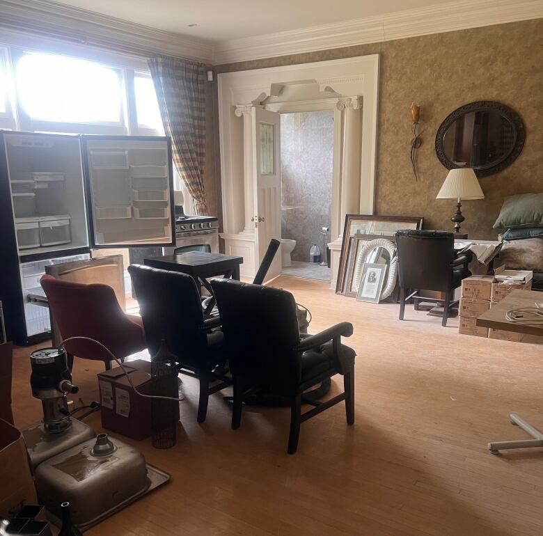 The interior of an abandoned victorian house. Chairs, a fridge and paintings sit on a messy floor.