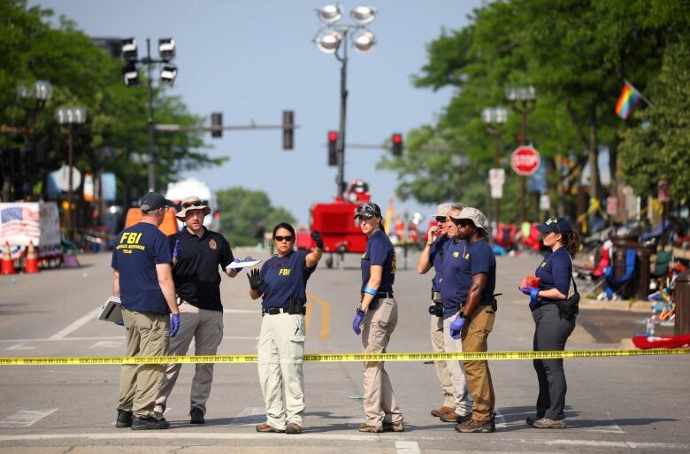 Eight law enforcement personnel stand on a street, along a strip of police tape.