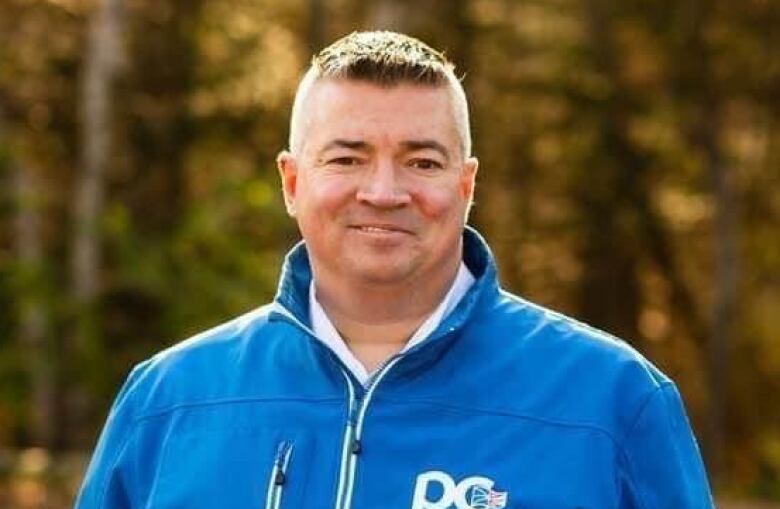 Man in blue coat with trees behind him.