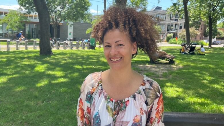 A smiling woman with curly hair tied in a ponytail sits on a bench in a park, wearing a flora top.