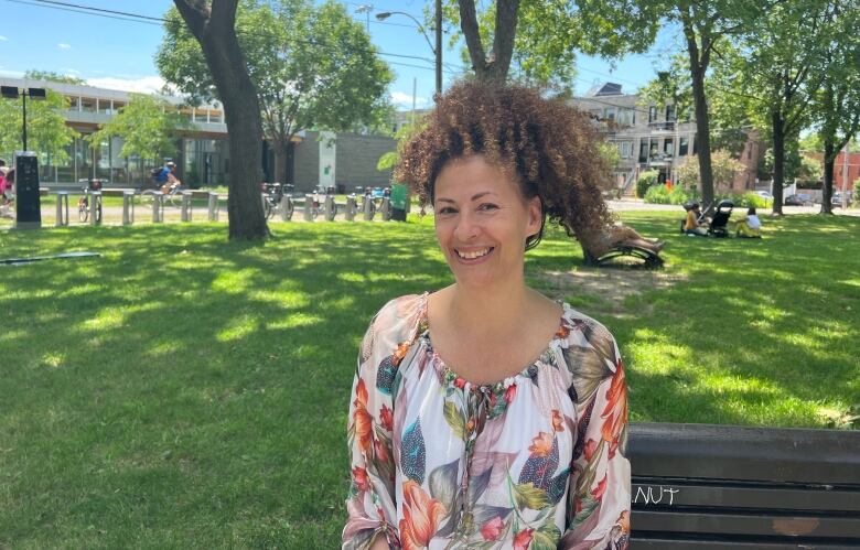 A smiling woman with curly hair tied in a ponytail sits on a bench in a park, wearing a flora top.