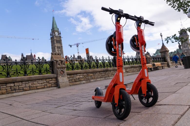 Two parked orange e-scooters in summer with a legislature in the background.