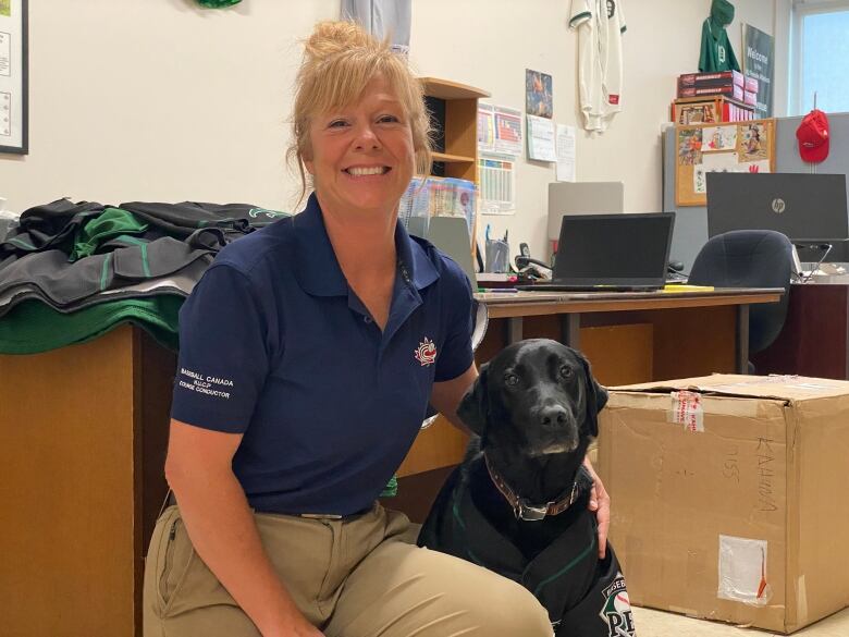 A smiling woman wearing blue polo shirt sits at an office desk with a large black labrador retriever by her side.