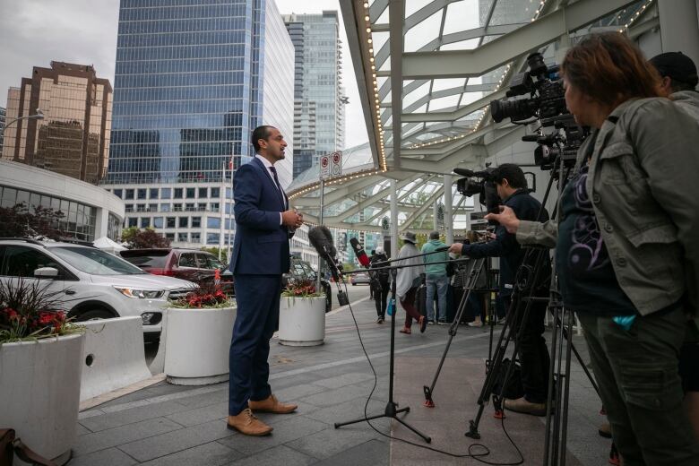 A South Asian man delivers a news conference in front of dozens of cameras in an outdoor environment.