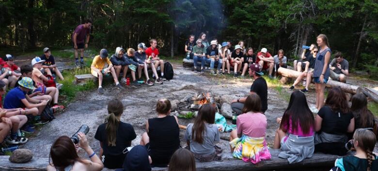 A camp counsellor stands to tell a story and approx. 50 kids gather around the bonfire to listen