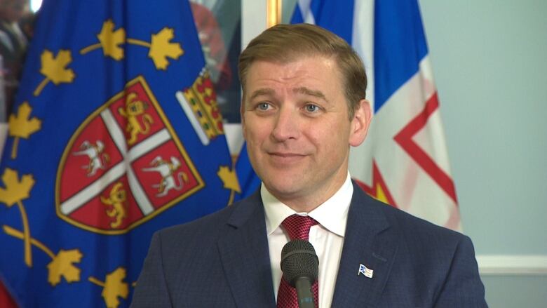 A man in a suit and tie stands in front of a microphone, with flags in the background.